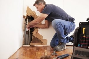 A plumber performs one of his many plumbing services on a pipe inside of a damaged wall.